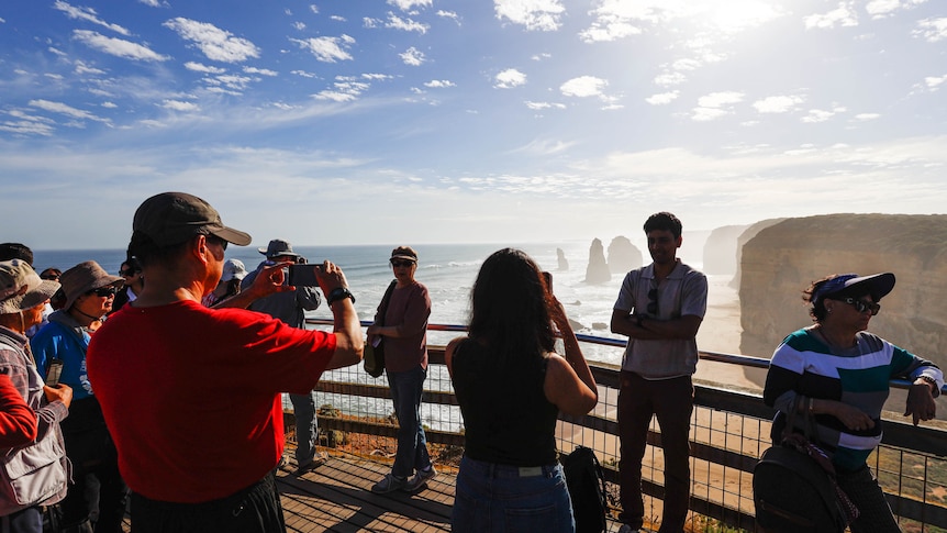 A massive crowd of tourists holding phones and cameras at the Twelve Apostles lookout in Victoria, Australia.