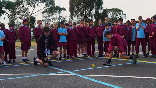Two kids playing a competitive game of handball or downball in a sunny school playground with a blue rubber ball.