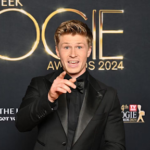 Television presenter Robert Irwin smiling broadly while wearing a sleek tuxedo on a glamorous red carpet.